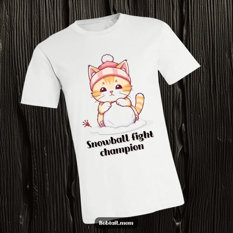 Funny T-shirt depicting a bobtail cat wearing a winter hat, diligently building a small snowball, ready for winter play.